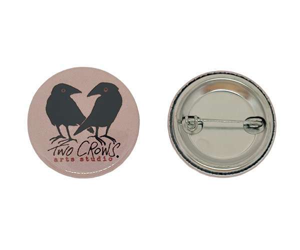 A 32mm Button Badge featuring two black crows facing each other and the text "Two Crows arts studio" on the front; the back displays the pin fastening mechanism.