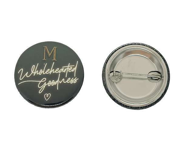 Two round black 32mm button badges. The front badge features "M Wholehearted Goodness" in gold and white script, whilst the back badge displays the metal pin clasp.
