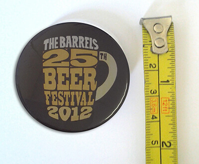 A round pin with "The Barrels 25th Beer Festival 2012" next to a yellow measuring tape showing a diameter of about 6 centimeters.