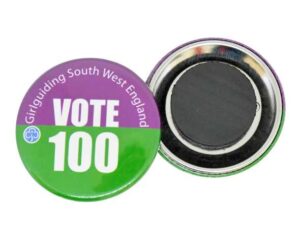 Round button badge with "Girlguiding South West England VOTE 100" written in white text on a purple and green background; the back of the badge is visible beside it.