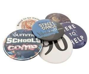 Six 77mm button badges with assorted round designs and texts, including "Street Dance," "Summer Schools Comp," "90," and "Here to Help," are displayed overlapping on a white background.