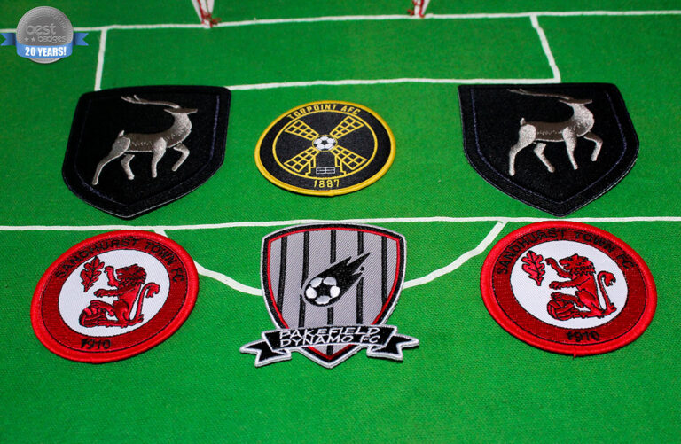 Six embroidered football club badges are arranged on a green field background, featuring various club logos and symbols, including a windmill, deer, and a dragon.