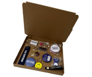 An open cardboard box displaying various keychains and round badges with different designs and texts arranged inside.