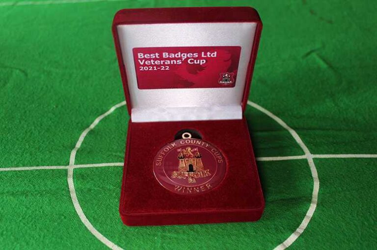 A medal in a red velvet box labeled "Best Badges Ltd Veterans’ Cup 2021-22 Winner" sits on a green surface marked like a soccer field.