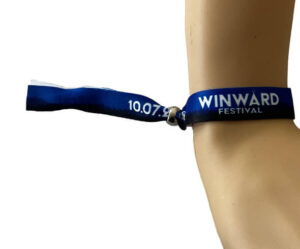 A blue fabric wristband with "WINWARD FESTIVAL" and "10.07" printed on it, worn on a person's wrist.