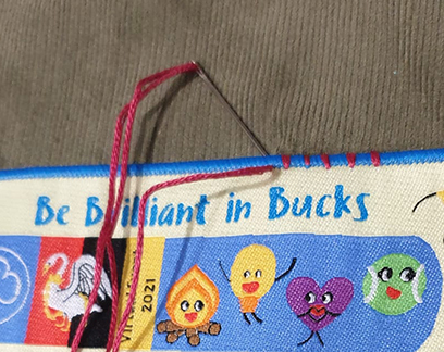 A needle with red thread is stitching the edge of a blue and yellow fabric strip that reads "Be Brilliant in Bucks" with cartoon faces below the text.