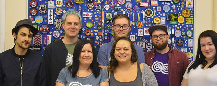 Seven people pose in front of a blue wall covered with assorted badges and patches, standing in two rows and facing the camera.