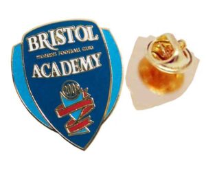 A blue and gold enamel pin with "Bristol Academy Women's Football Club" text, next to a gold pin backing.
