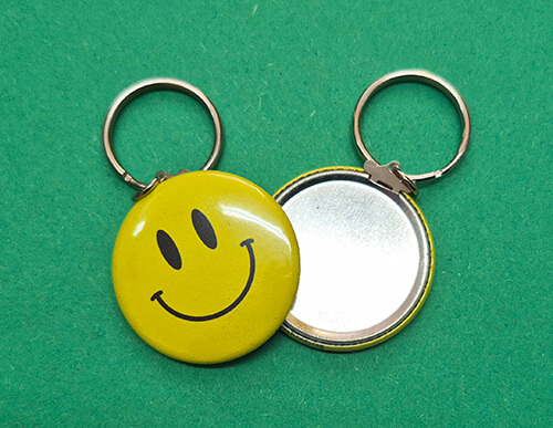 Two round keychains on a green surface; one features a yellow smiley face, the other is open and shows a blank metallic surface.
