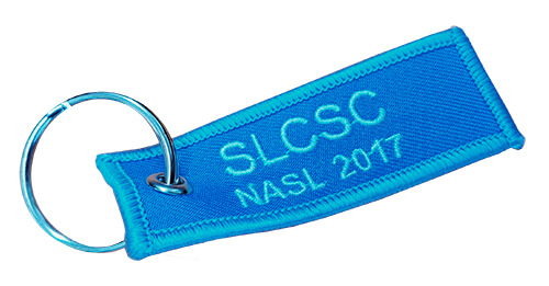 A blue fabric keychain with "SLCSC NASL 2017" embroidered in light blue thread, attached to a metal key ring.