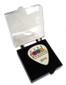 A white guitar pick labeled "Krisey and Friends Music Making Memories 2015" displayed in an open black and clear plastic case.