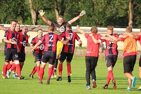 A soccer team in red and black uniforms celebrates together on a grass field, with one player jumping in the air with arms raised.