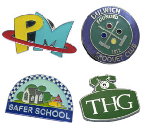 Four colorful enamel pins: one with "PM" and a red ring, one for Dulwich Croquet Club, one for Safer School with a house, and one green pin labeled "THG" with a telephone icon.