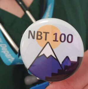 A person wearing a lanyard and stethoscope holds a round pin featuring "NBT 100" with a mountain and sun design.