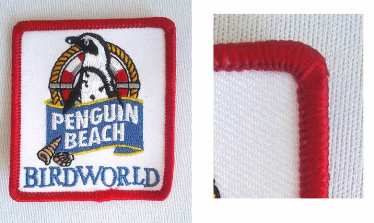 A rectangular embroidered patch with a red border features a penguin, a lifebuoy, and the text "PENGUIN BEACH BIRDWORLD" on a white background.