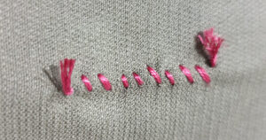 Pink embroidery thread is sewn in a zigzag pattern through grey fabric, with frayed ends visible on both sides.