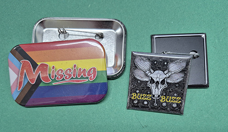 Two pins on a green surface: one with a rainbow "Missing" design, and another with a skull and bee wings labeled "Buzz Buzz," both showing their pin backs.