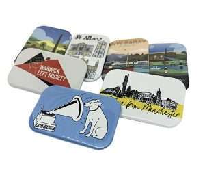 Seven 70mm x 45mm rectangular button badges, featuring various colourful illustrations and designs—including landmarks and a dog listening to a gramophone—are arranged in a loose cluster on a white background.