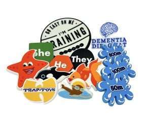 A set of Soft PVC Badges showcases a variety of colourful designs, including animals, pronouns, water splashes, and text like “I’M TRAINING” and “DEMENTIA DINOSAUR.”.