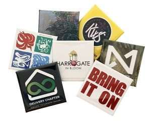 Seven 38mm Square Button Badges with various colourful logos and text, including "BRING IT ON" and "HARROGATE IN BLOOM," arranged in an overlapping group on a white background.