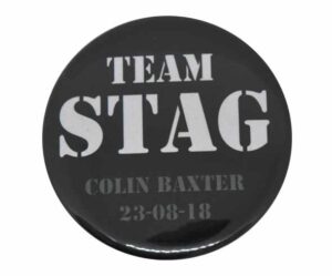 A black button badge with "TEAM STAG" in large white letters and "COLIN BAXTER 23-08-18" in smaller gray text below.