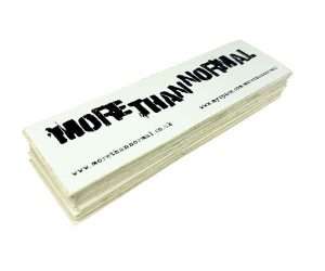 A batch of 1,000 rectangular white vinyl stickers featuring the text “MORE-THAN-NORMAL” in bold, distressed black font and website addresses printed underneath.