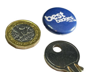 A one pound coin, a blue pin badge with "bestbadges.co.uk" in white text, and a metal key on a white background.