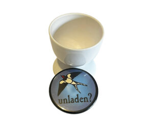 A white egg cup next to a round pin featuring a flying swallow and the word "unladen?" on a blue background.