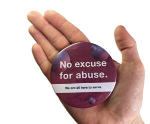 A hand holds a round badge that reads, "No excuse for abuse. We are all here to serve." The badge has a purple background with dark circular patterns.
