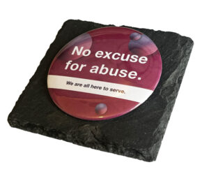 A round sign on a black slate reads "No excuse for abuse. We are all here to serve." in white and yellow text on a maroon background with circular patterns.
