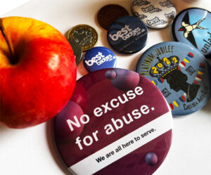 An apple, coins, and various badges are arranged on a white surface. The largest badge reads, "No excuse for abuse. We are all here to serve.