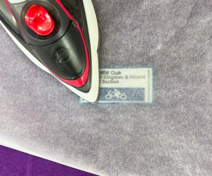 A close-up of an iron pressing over parchment paper, transferring a label with text and a bicycle graphic onto fabric underneath.