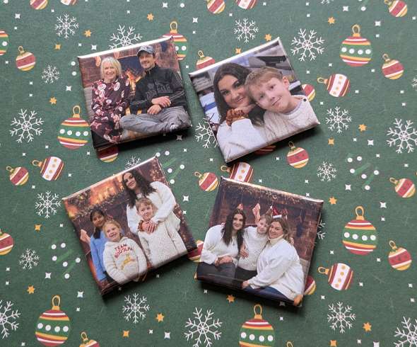 Four square photo magnets featuring family portraits are arranged on green holiday-themed wrapping paper with ornament and snowflake patterns.