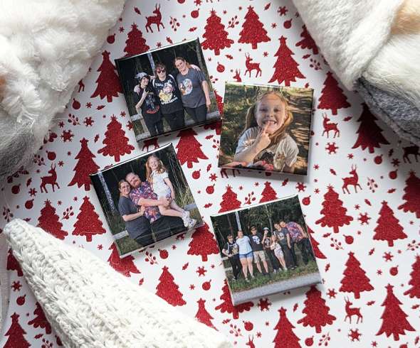 Four square family photo magnets are arranged on festive red and white Christmas-themed paper, surrounded by soft knit and fleece fabrics.