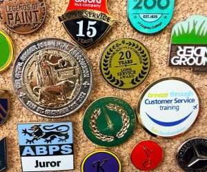 Assorted pins and badges, including enamel badges for long service awards, a customer service training badge, a juror badge, and decorative emblems, displayed on a cork board.