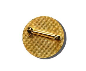 A gold-colored brooch pin with a horizontal clasp is attached to a textured circular fabric background.